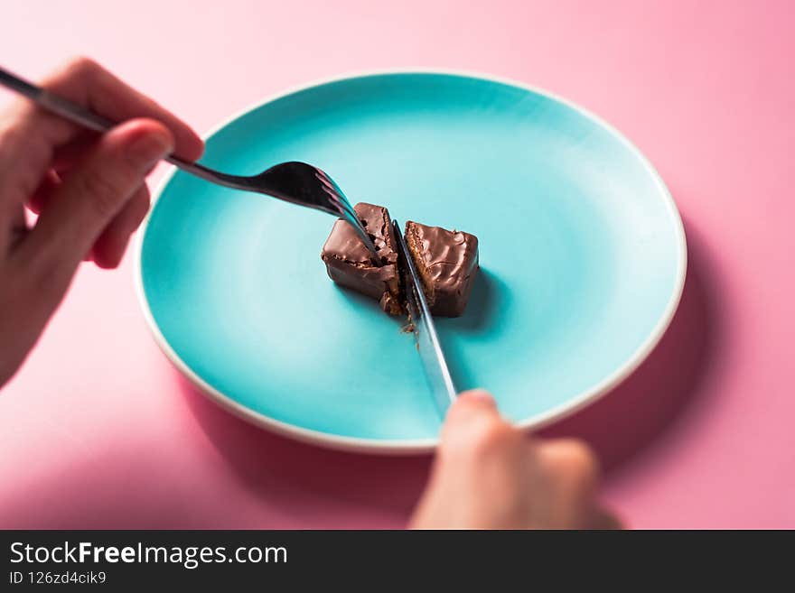 Cropped view of woman hands cutting dessert chocolate bar with knife and fork on colored plate on pink background. Cropped view of woman hands cutting dessert chocolate bar with knife and fork on colored plate on pink background.
