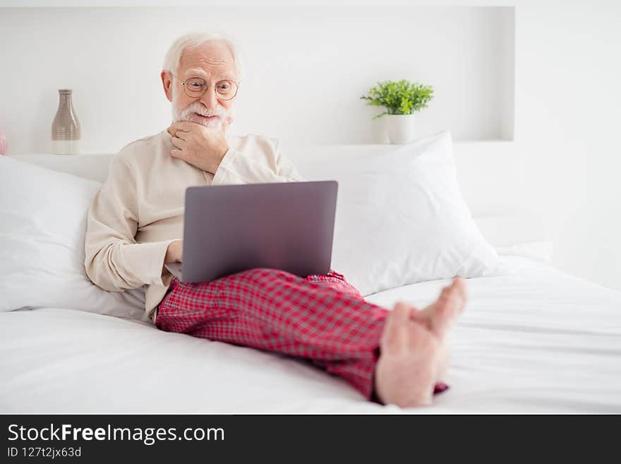 Photo of aged man pensioner serious think look browse read laptop internet online lying bed hand touch chin indoors.