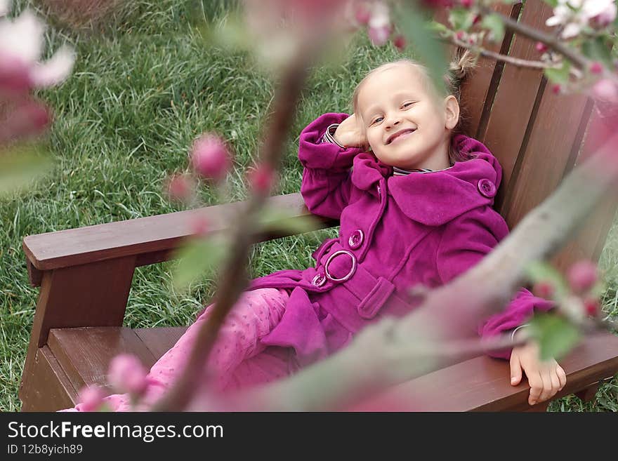 Spring garden baby. Little girl in spring garden near flowers. Child in purple coat sits on wooden chair in garden