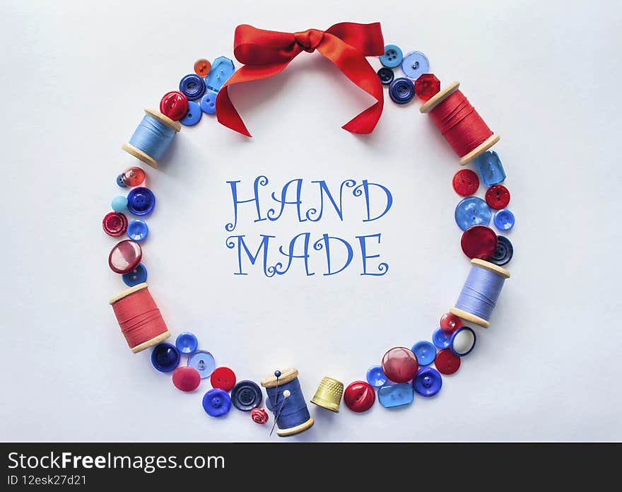 The wreath is made of buttons and threads of red and blue colors with pins and a bright bow of satin ribbon at the top. On a white background. The concept of craft, sewing and embroidery. In the middle is an inscription Handmade.