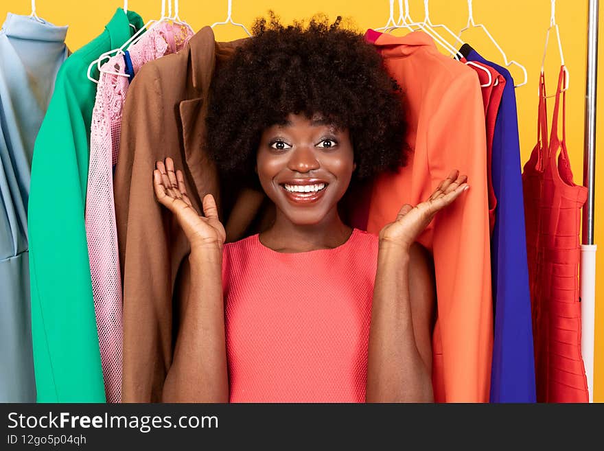 Happy African Female Shopping Posing Among Colorful Clothes, Yellow Background