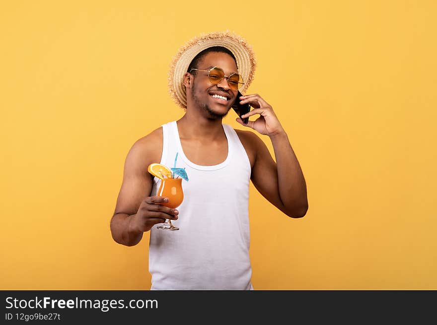I&#x27;m on vacation. Positive black guy in trendy outfit holding tropical fruit cocktail and speaking on smartphone on yellow studio background. African American man enjoying his summer holidays. I&#x27;m on vacation. Positive black guy in trendy outfit holding tropical fruit cocktail and speaking on smartphone on yellow studio background. African American man enjoying his summer holidays
