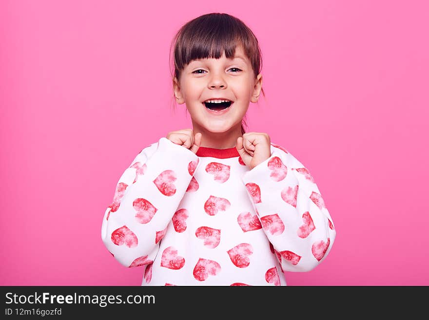 Happy excited little girl clenching fists, celebrating her success, looking at camera with excited emotions, cant believe she is winner, isolated over pink background