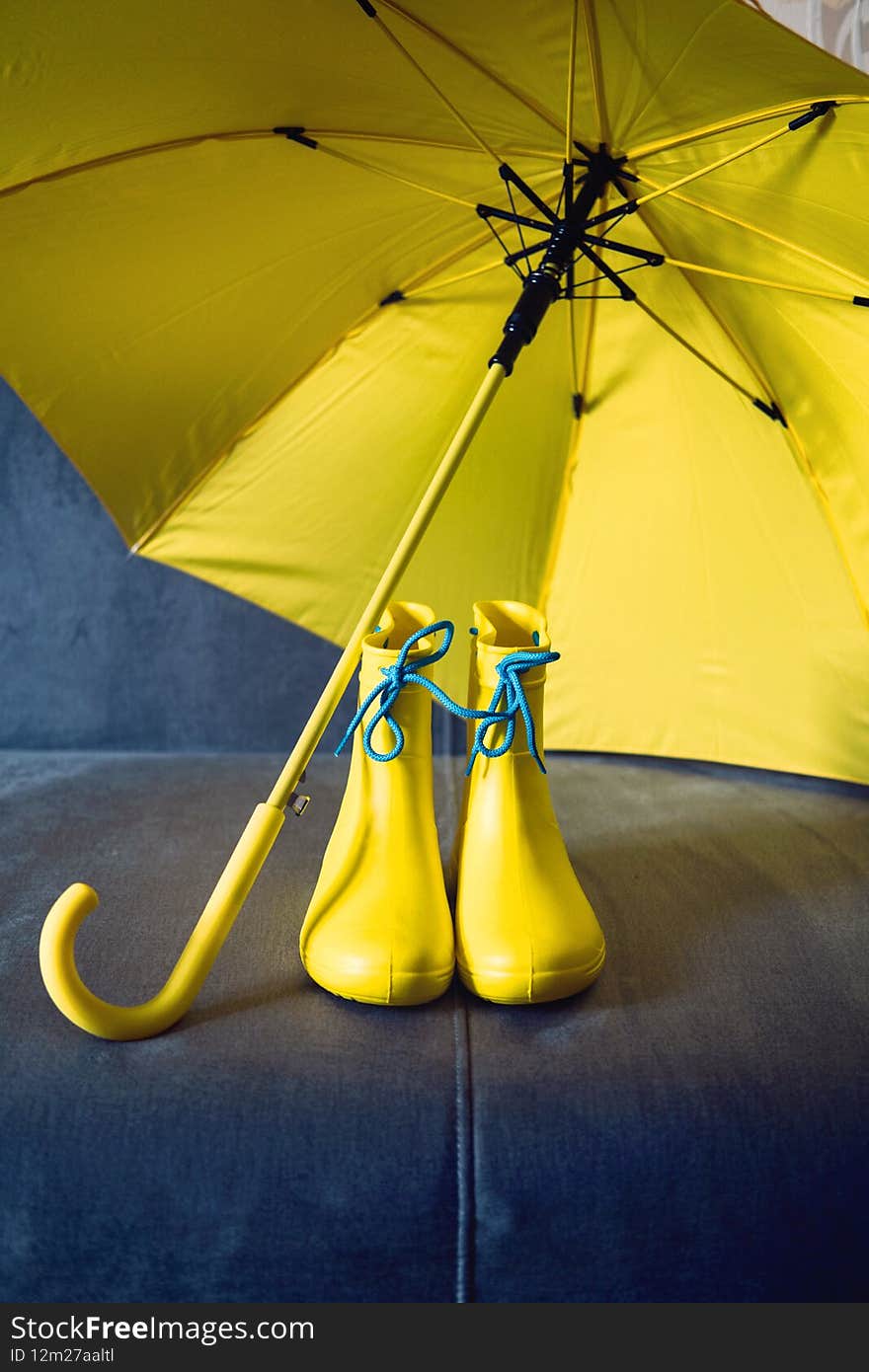 Rainy Mood, outfit for rainy day. Blue raincoat, yellow rubber boots and umbrella on a gray sofa. Colors of the year Ultimate Gray and Illuminating yellow background.