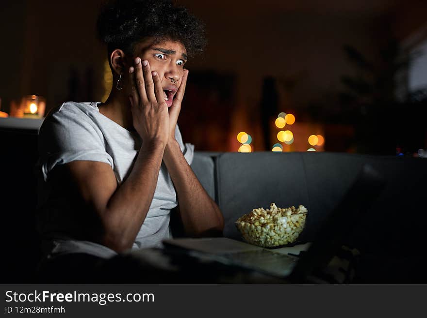 Young guy looking terrified while watching horror movie alone using laptop and eating popcorn, sitting on a comfortable sofa in dark room at home. Evening routine, leisure concept