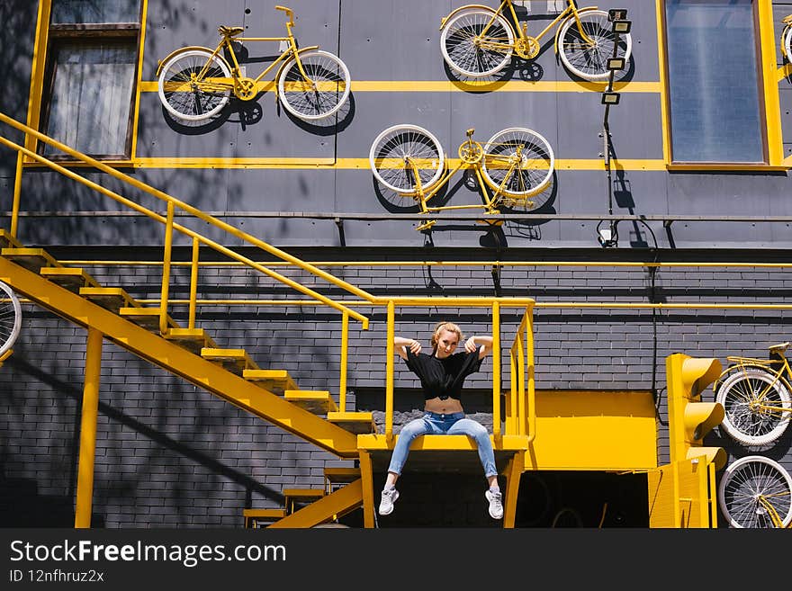 A young girl is sitting on the stairs against the background of a creative building. The gray wall is decorated with yellow bicycles.Colors of the year 2021. Creative approach to construction
