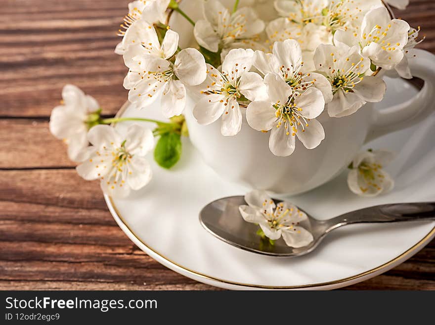 White porcelain cup in it flowers are white from the apple tree. On a brown background. Spoon on a platter.