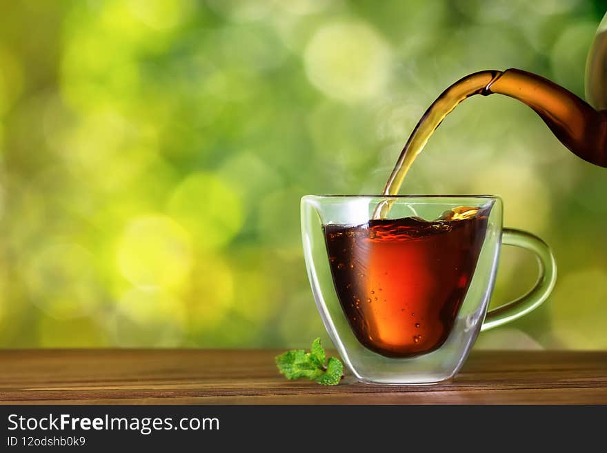 black tea with mint flavor pouring from glass teapot into double sided wall cup on wooden table with green natural blurred background. black tea with mint flavor pouring from glass teapot into double sided wall cup on wooden table with green natural blurred background