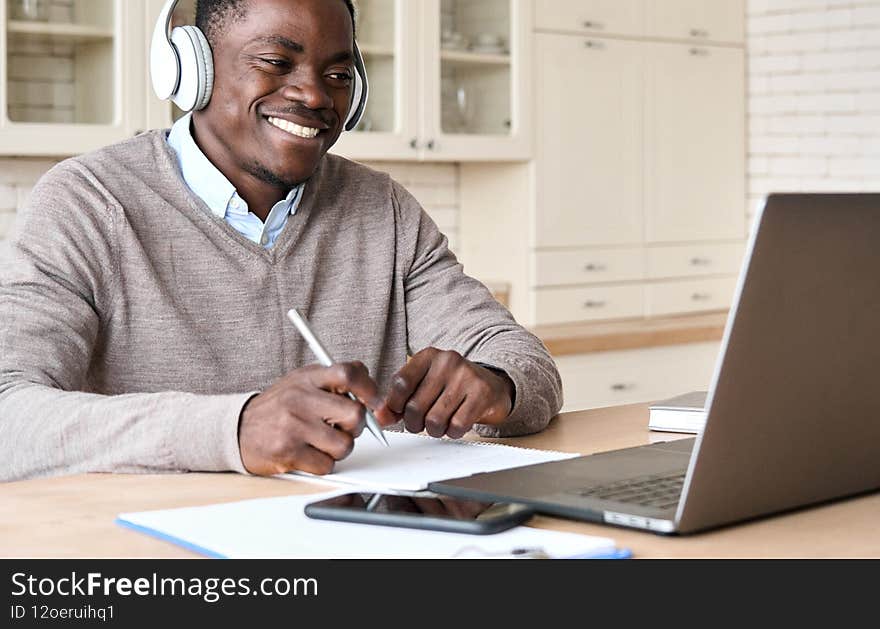 Adult happy smiling African American black male student wearing headphone sit at kitchen table, watching online video conference learning virtual course teaching using laptop computer in homeoffice. Adult happy smiling African American black male student wearing headphone sit at kitchen table, watching online video conference learning virtual course teaching using laptop computer in homeoffice.