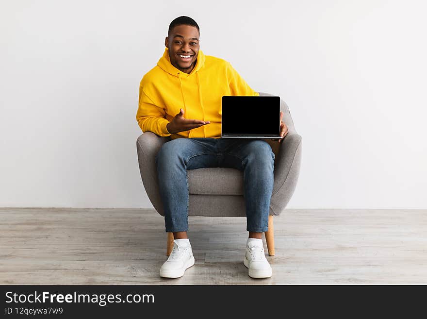 Great Website. African American Man Showing Laptop Computer With Blank Screen Recommending Online Service Pointing Finger At Display Sitting In Chair Over Gray Wall Background. Mockup. Great Website. African American Man Showing Laptop Computer With Blank Screen Recommending Online Service Pointing Finger At Display Sitting In Chair Over Gray Wall Background. Mockup