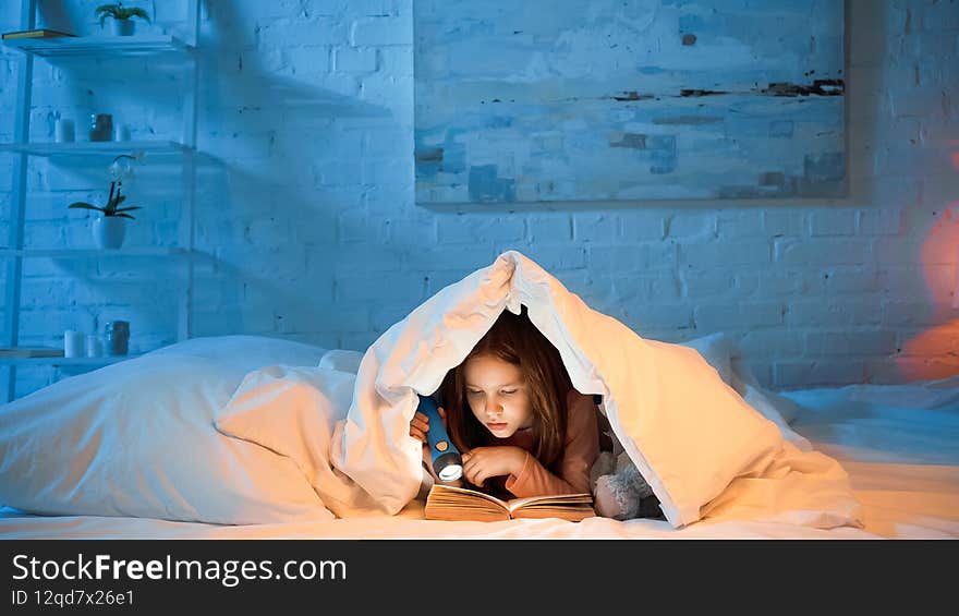 Child under blanker reading book and holding flashlight,stock image