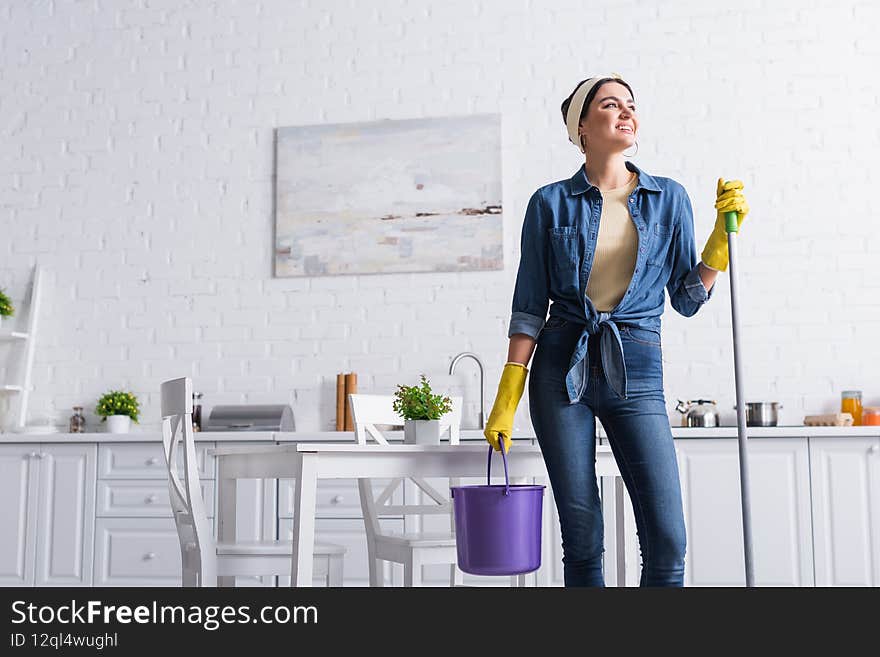 Cheerful woman in rubber gloves holding mop and bucket in kitchen,stock image