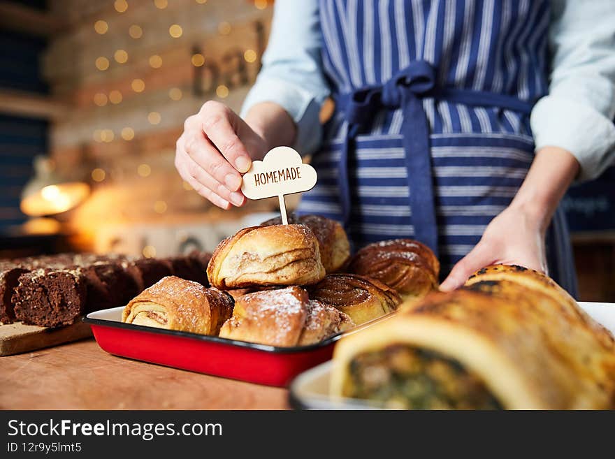 Close Up Of Sales Assistant In Bakery Putting Homemade Label Into Stack Of Freshly Baked Baked Cinnamon Buns