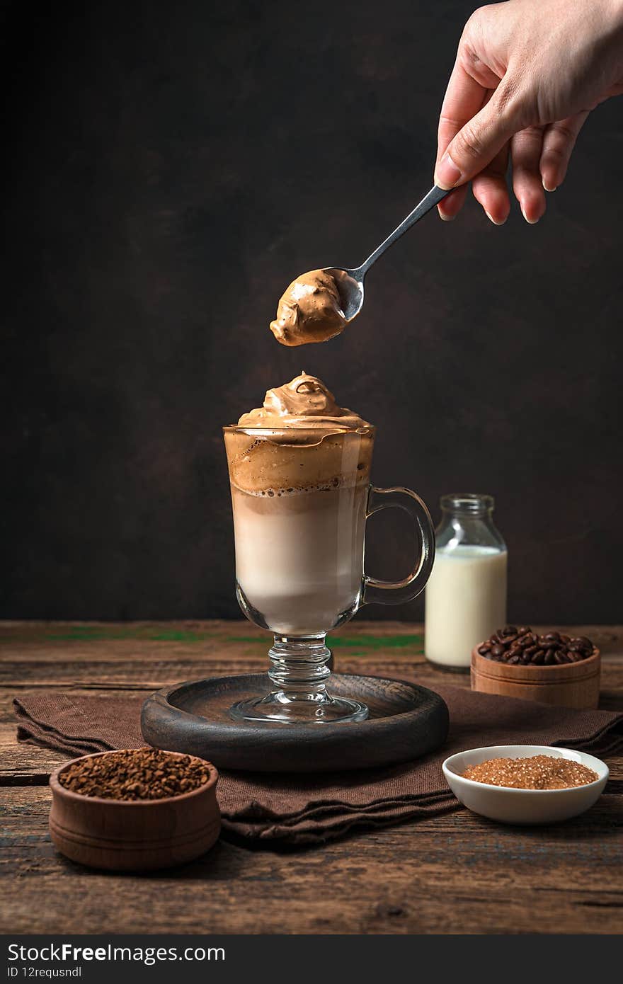 A woman& x27;s hand puts coffee foam in a glass of dalgona coffee on a brown background.