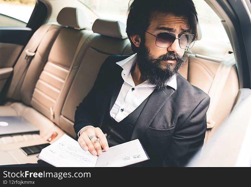 A stylish and successful Indian with glasses and a beard works and plans his schedule in the back seat of the car. Safe and comfortable business travel,