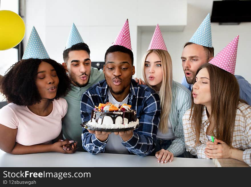 Cheerful multiethnic friends blowing lit candles on birthday cake, wearing party hats, enjoying festive occasion. Happy diverse millennials celebrating b-day together at home. Cheerful multiethnic friends blowing lit candles on birthday cake, wearing party hats, enjoying festive occasion. Happy diverse millennials celebrating b-day together at home