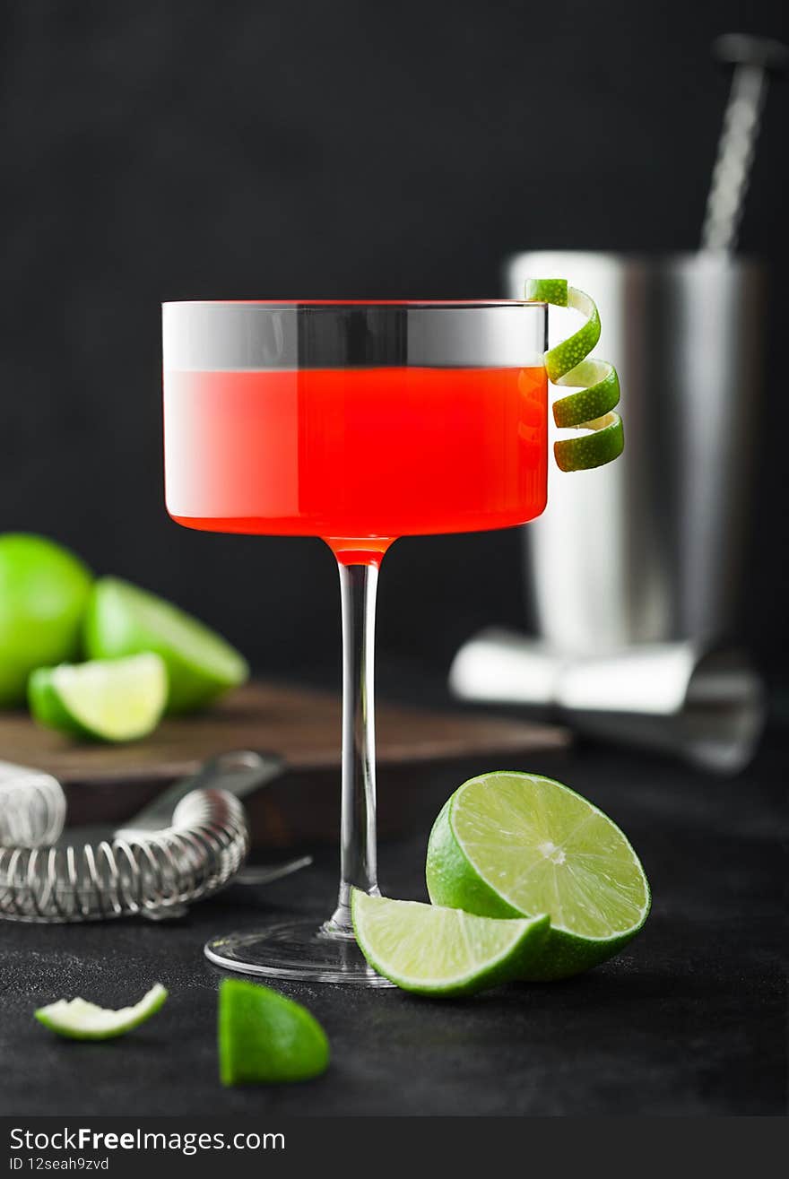 Cosmopolitan cocktail in modern crystal glass with lime peel and fresh limes with strainer, jigger and shaker on black table background