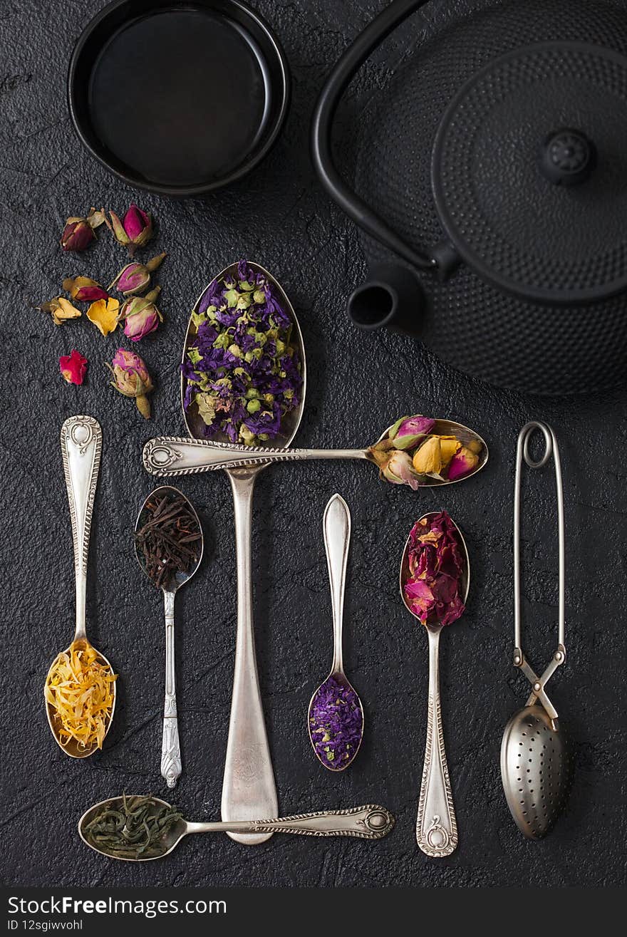 Spoons with various classic and herbal tea on black background with iron teapot and cup with vintage strainer infuser. Top view. Spoons with various classic and herbal tea on black background with iron teapot and cup with vintage strainer infuser. Top view