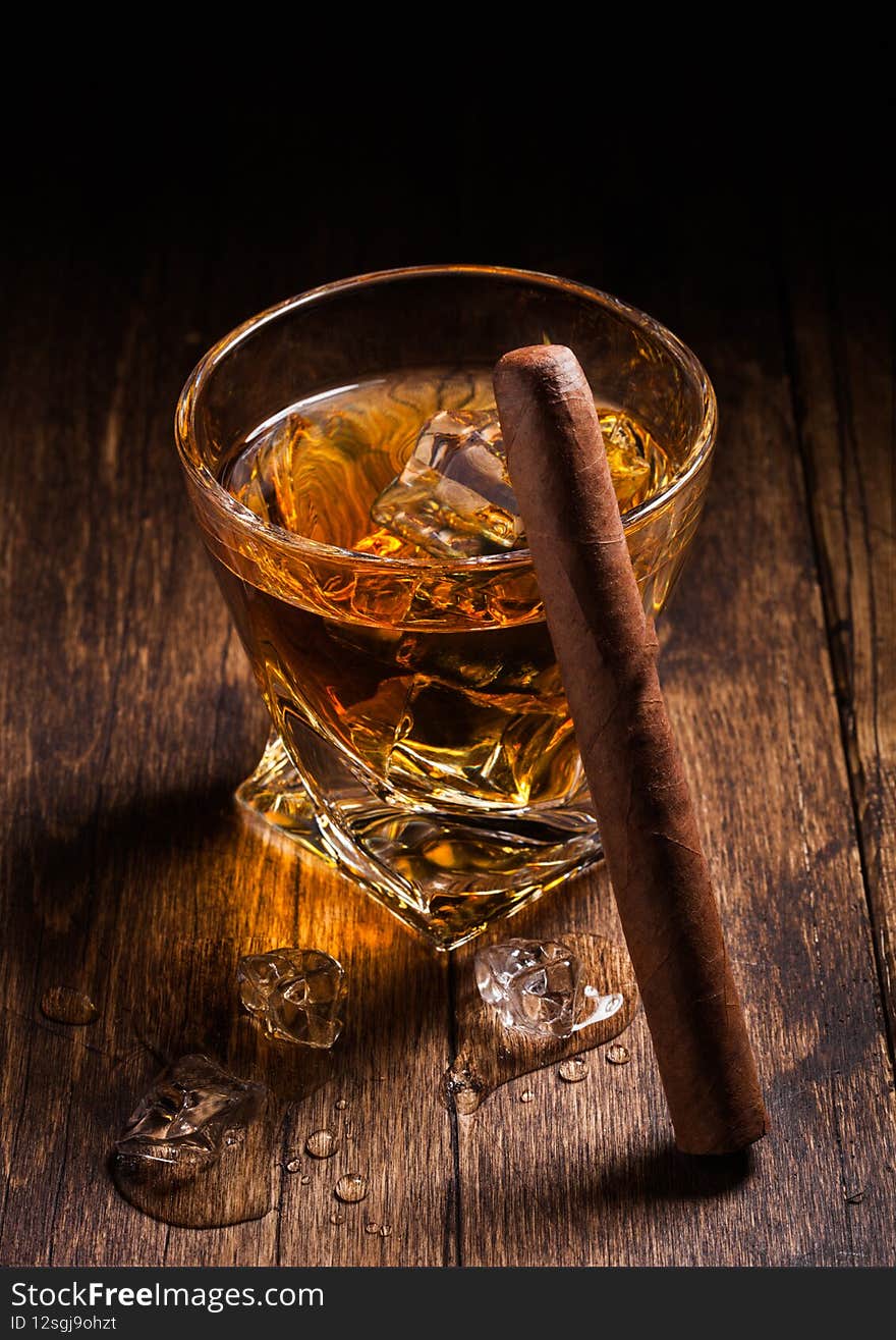 Single malt scotch whiskey in modern crystal glass with ice cubes and cuban cigar on wooden background