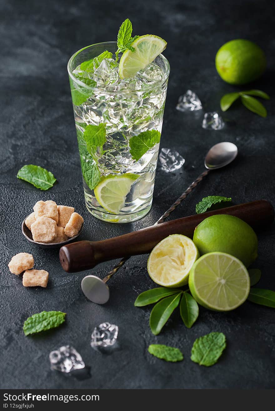 Glass of Mojito cocktail with ice cubes mint and lime on black board with spoon and muddler and fresh limes and cane sugar.