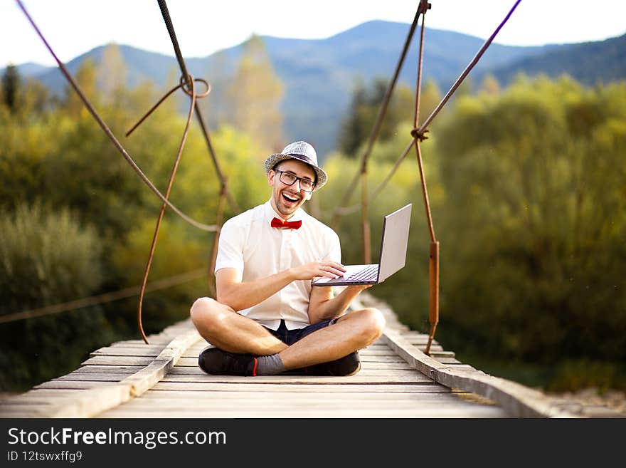 Intelligent handsome male in hat, white shirt and glasses sit with crossed legs and holding a laptop. Man working outdoors with laptop sitting in mountains forest. Concept of remote work and freelancer lifestyle. Intelligent handsome male in hat, white shirt and glasses sit with crossed legs and holding a laptop. Man working outdoors with laptop sitting in mountains forest. Concept of remote work and freelancer lifestyle.