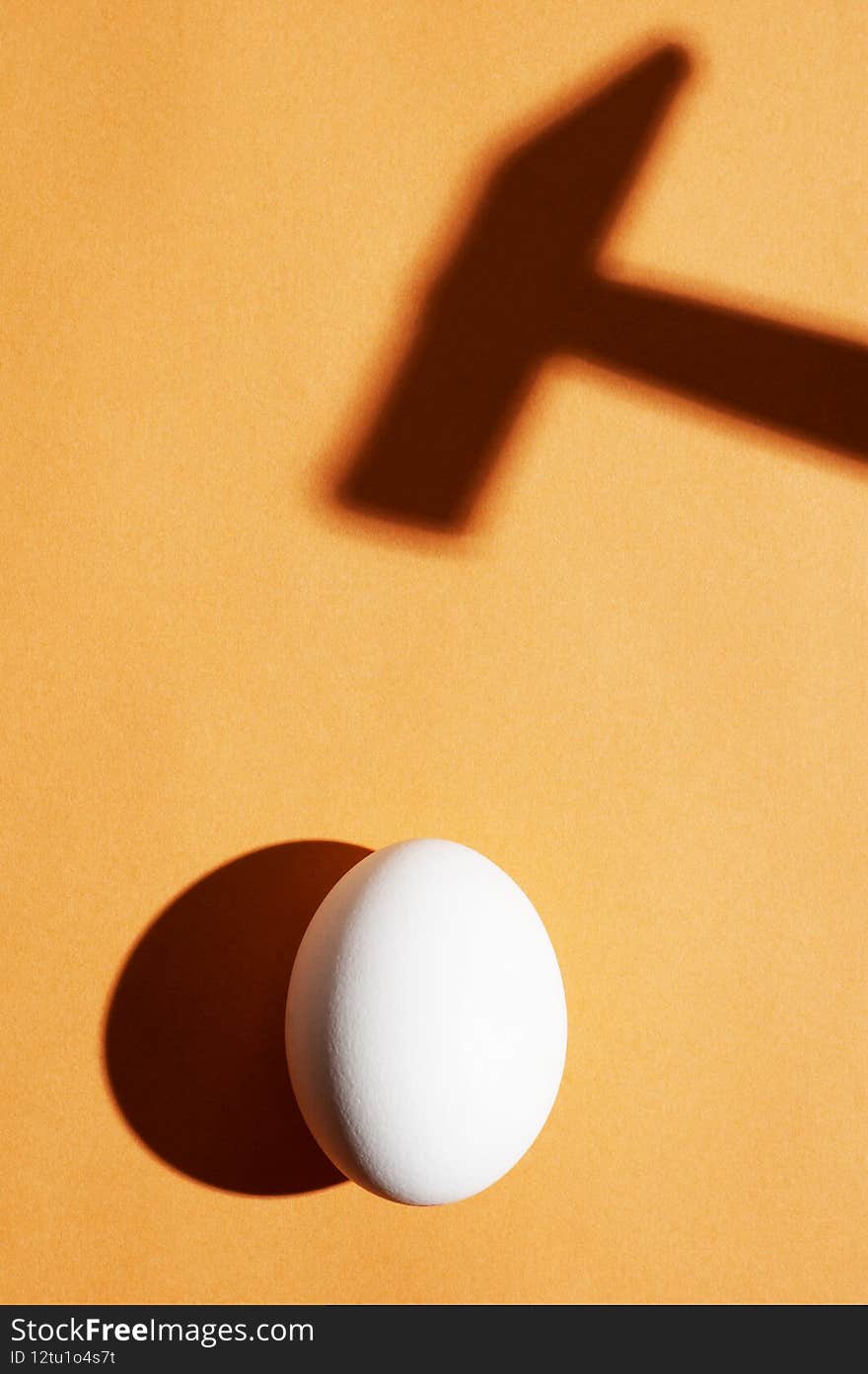Minimal concept with an egg under the harsh light and a hammer shadow on an orange background