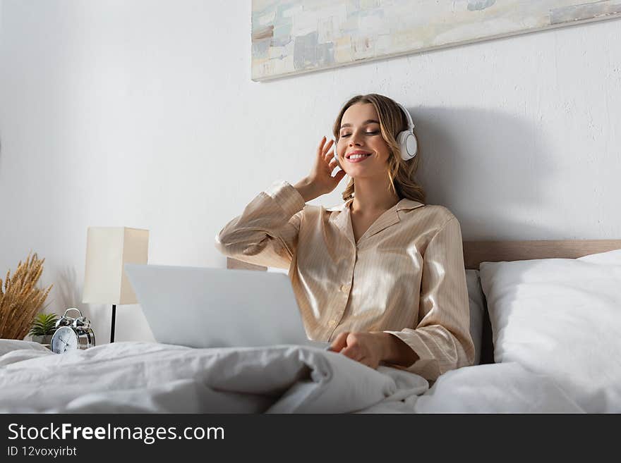 Cheerful woman listening music in headphones near blurred laptop on bed,stock image
