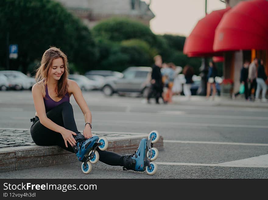 Pleased dark haired European woman puts on inline skates going rollerblading poses against blurred city background