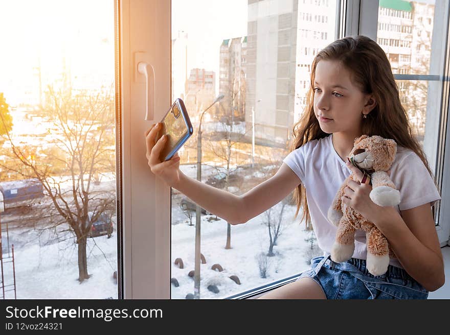 cute teenage girl sits on the windowsill with a stuffed toy and a phone and takes a selfie. The sun is shining outside the window