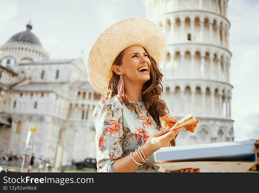 Smiling trendy woman in floral dress with pizza and hat near Leaning Tower in Pisa, Italy
