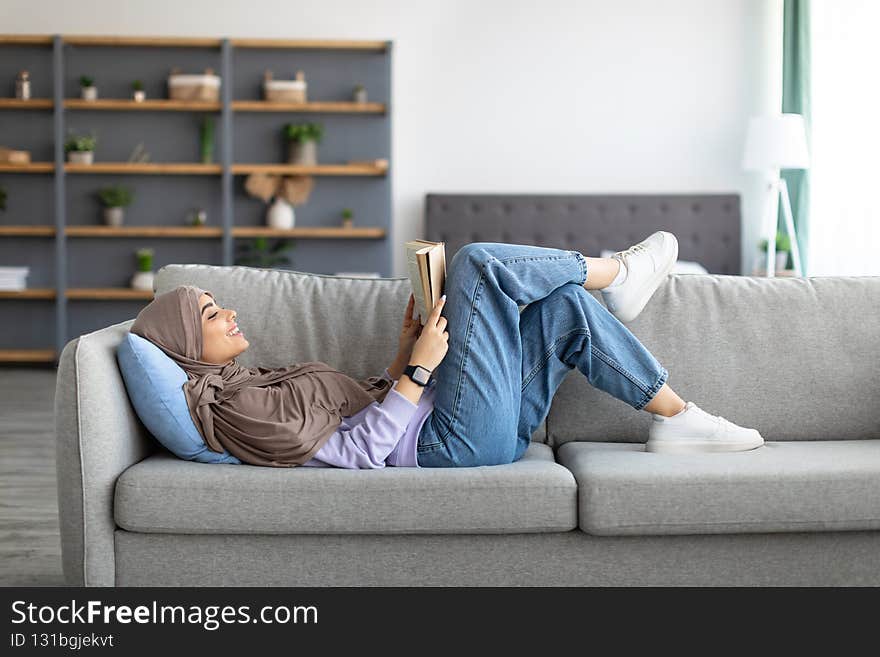 Free Time To Relax. Profile of beautiful muslim woman lying on comfortable couch at home, reading paper book. Happy lady in headscarf taking break, resting in living room, free copy space. Free Time To Relax. Profile of beautiful muslim woman lying on comfortable couch at home, reading paper book. Happy lady in headscarf taking break, resting in living room, free copy space