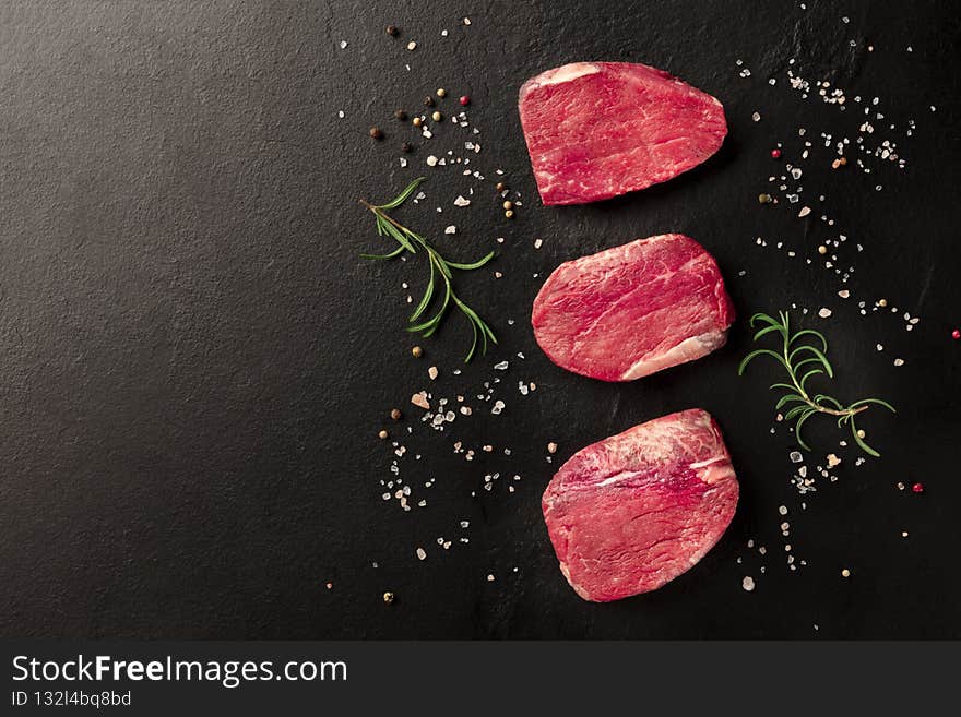 Raw beef steaks with salt, pepper and rosemary, shot from above on a black background with copyspace