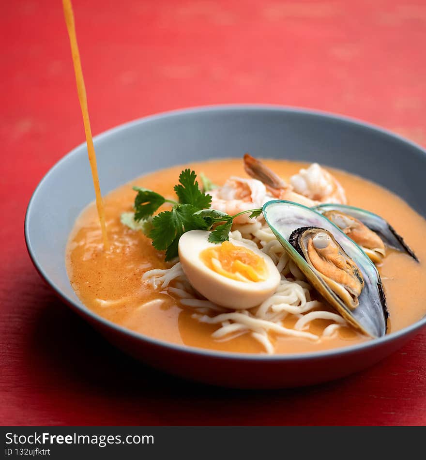 Broth pouring in Ramen soup. Bowl of noodles with mussels or seafood and egg on red background. Spicy oriental dish, Pan-Asian cuisine. Close up shot.