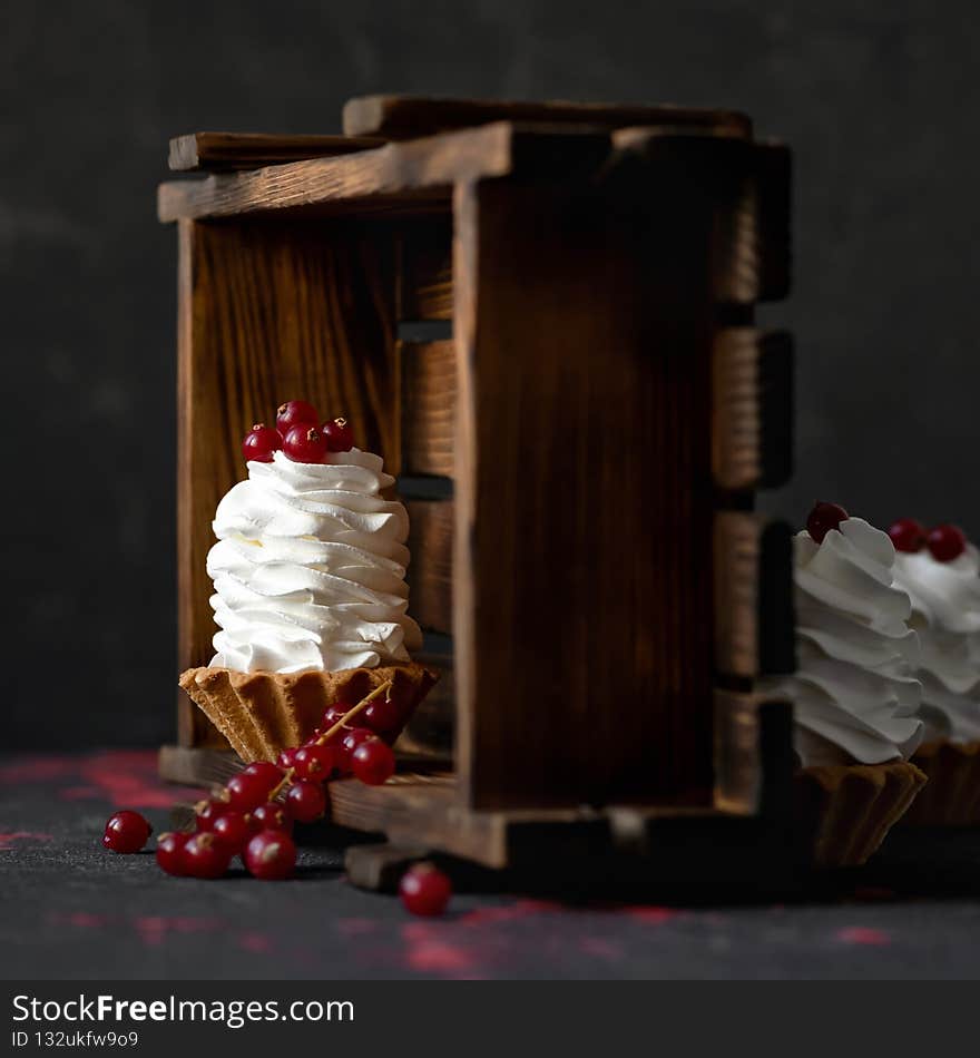 Puffy Creamy dessert decorated with red berries. Baked tarts or baskets of dough with airy whipped cream or mousse on dark background with wooden box. Mini cake. Copy space.