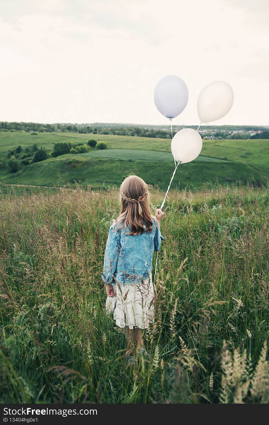 Kid stands along in grass on hilltop, looks into distance, holds three balloons. Girl walks in open air. Childhood, freedom, enjoy