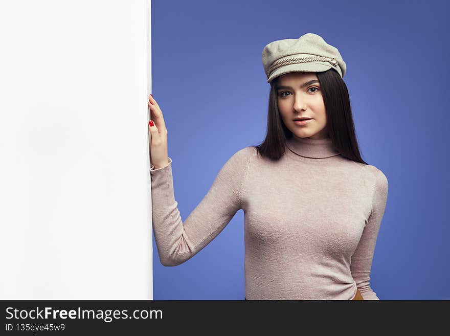 Portrait of charming lady standing alongside empty white billboard wall. Pretty girl wearing violet sweater, isolated over purple background. Portrait of charming lady standing alongside empty white billboard wall. Pretty girl wearing violet sweater, isolated over purple background