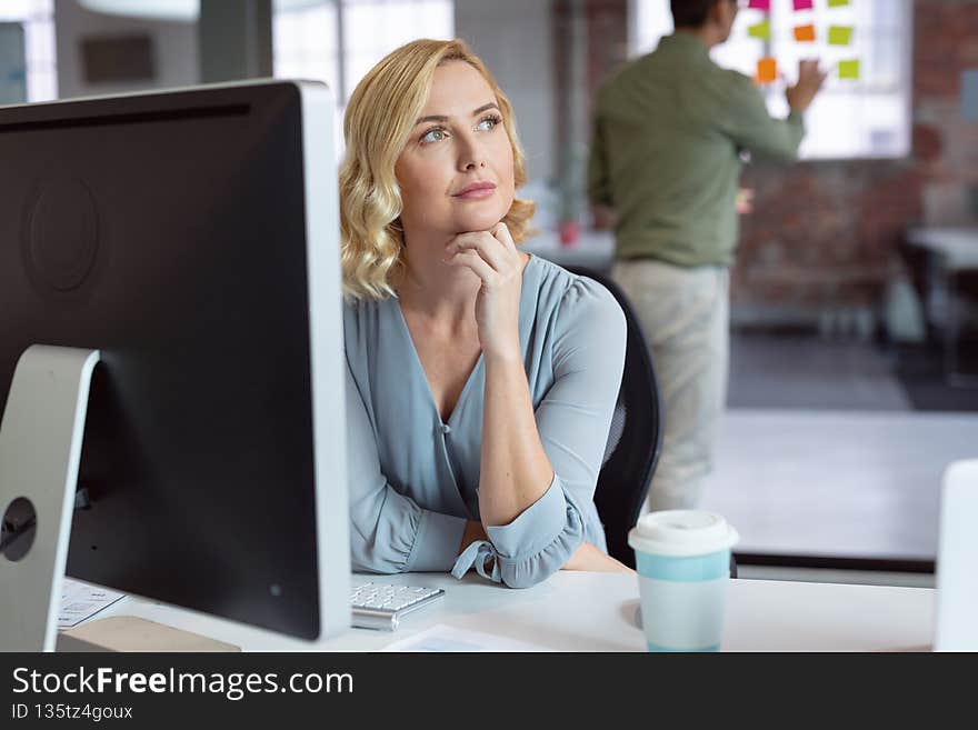 Thoughtful caucasian businesswoman sitting at desk using computer with male colleague in background