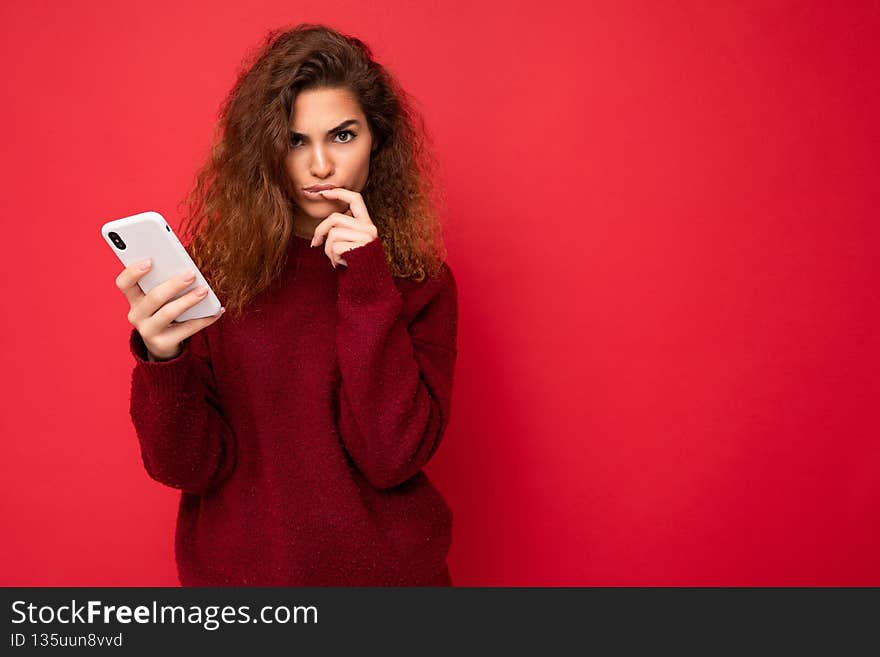 Photo of pretty young female person with curly hair wearing dark red sweater isolated on red background holding mobile phone looking at camera and thinking keeping finger near lips. Copy space