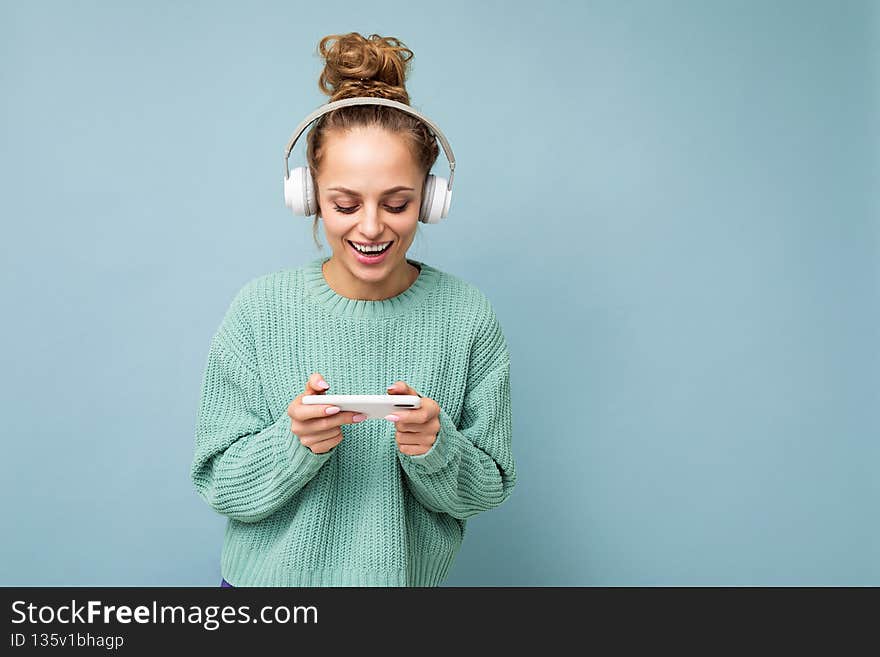 Beautiful positive smiling young woman wearing blue sweater isolated over blue background wearing white bluetooth
