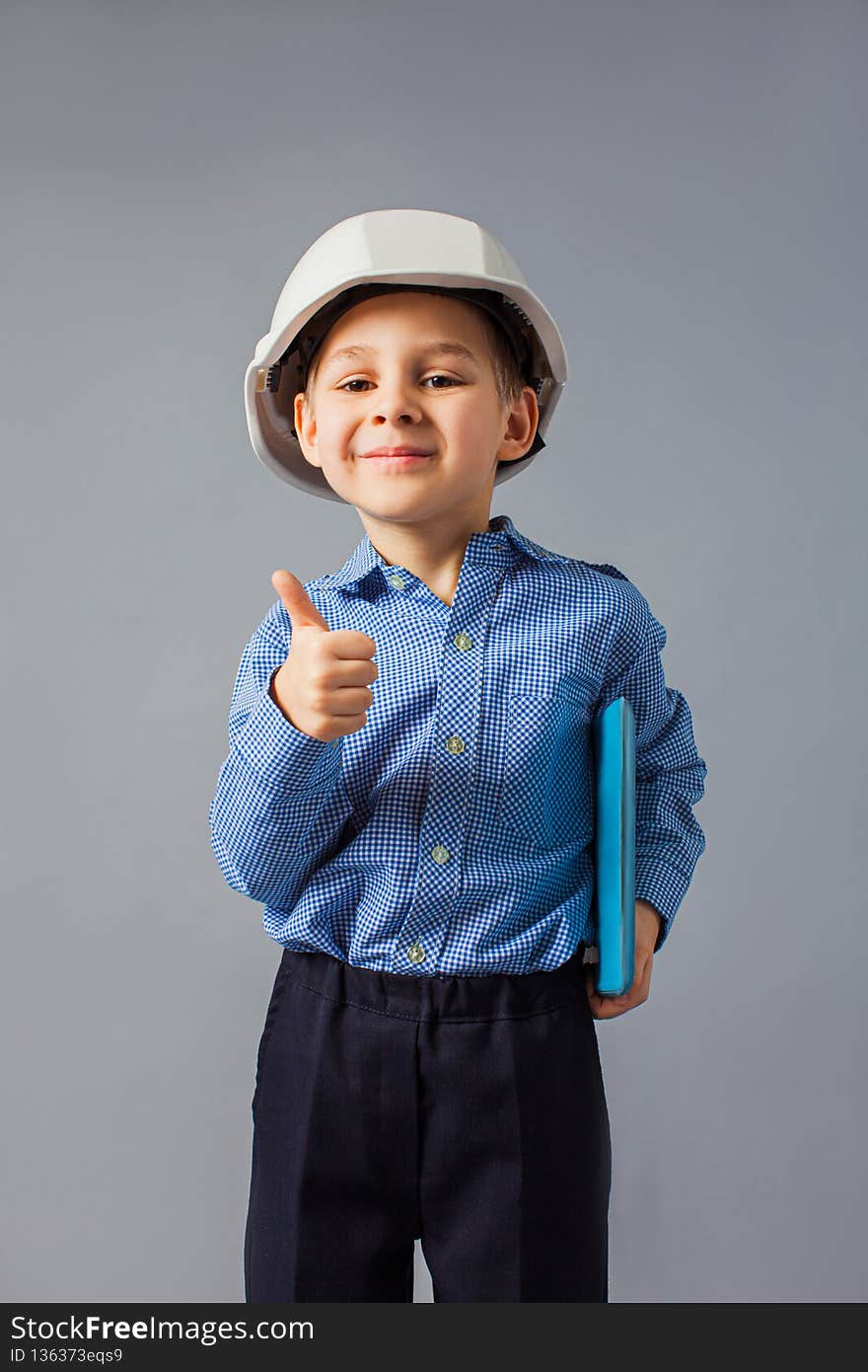 The little boy dressed in a costume of engineer stay at the gray background. The happy boy in a white helmet is looking on the tablet. The little boy dressed in a costume of engineer stay at the gray background. The happy boy in a white helmet is looking on the tablet
