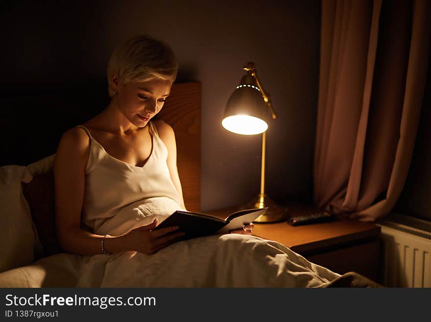 Beautiful female in pajamas lying on bed reading book, alone at night