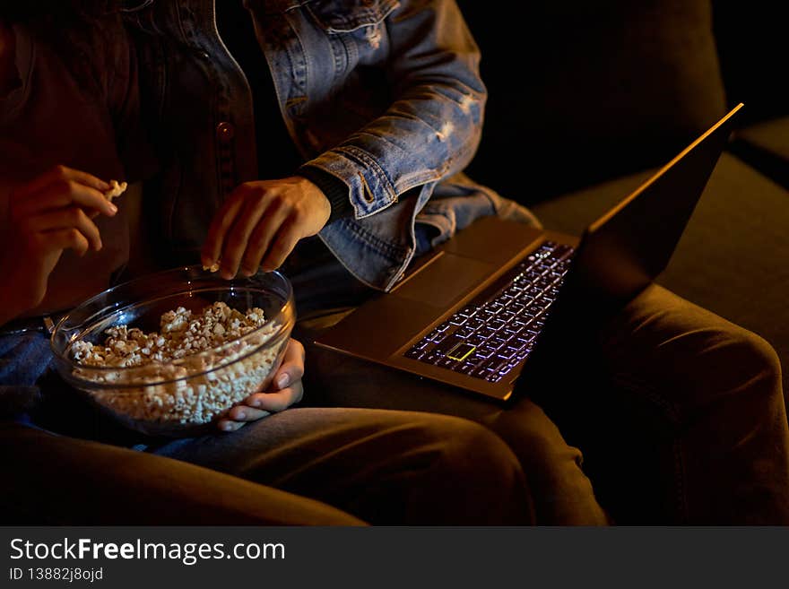 Cropped couple watching movie on laptop, eating popcorn at night cozy home