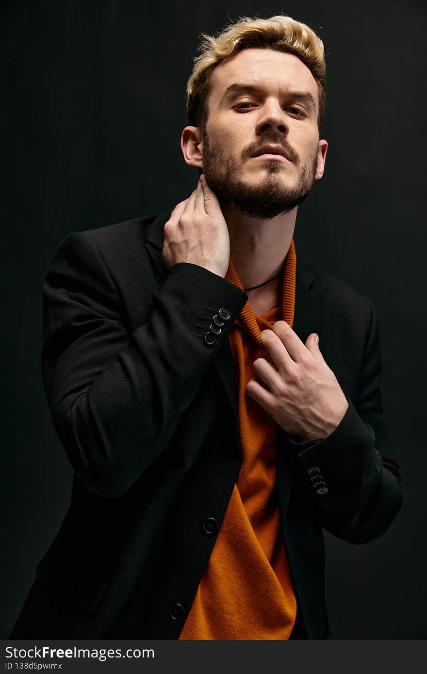 a blond man in a sweater and a jacket touches his neck on a dark background. High quality photo
