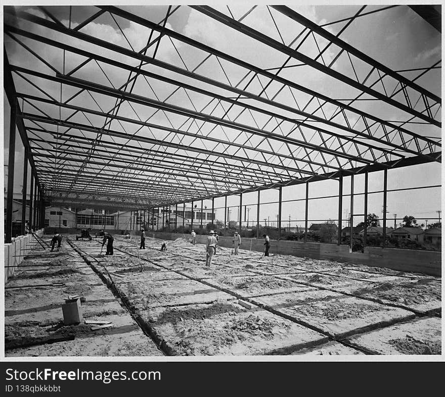 Photograph of a warehouse and office building during construction. The building was constructed by Alco Products, Inc. out of Beaumont using materials provided by John Dollinger Jr., Inc. The steel framing has been completed and workers appear to be sweeping the area, which appears to be covered with dirt. Houses and a large building are in the background. Photograph of a warehouse and office building during construction. The building was constructed by Alco Products, Inc. out of Beaumont using materials provided by John Dollinger Jr., Inc. The steel framing has been completed and workers appear to be sweeping the area, which appears to be covered with dirt. Houses and a large building are in the background.