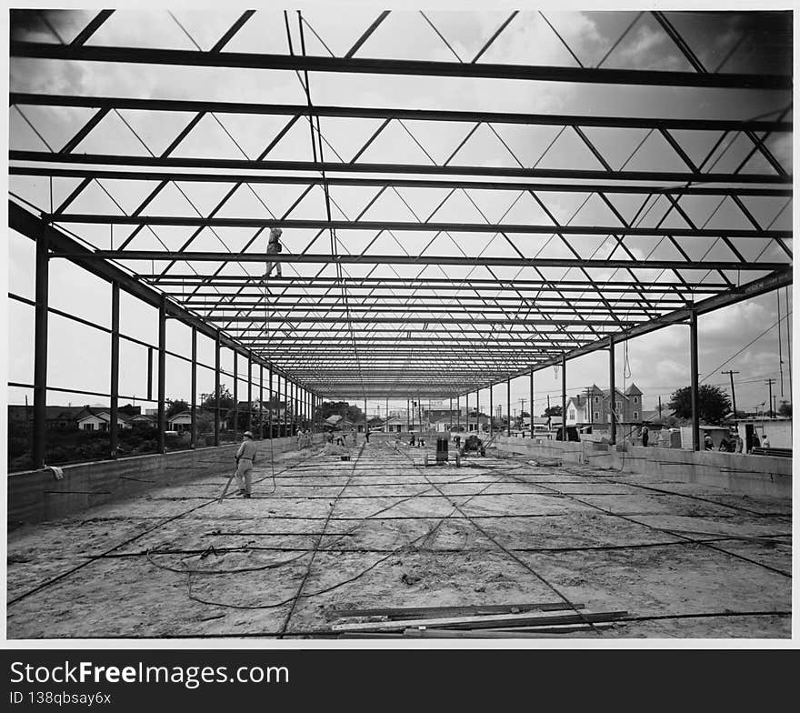 Photograph of a warehouse and office building during construction. The building was constructed by Alco Products, Inc. out of Beaumont using materials provided by John Dollinger Jr., Inc. The steel framing has been completed and workers appear to be sweeping the area, which is a dirt floor. An additional man is walking across a beam at the top of the photograph. Houses and a large building are in the background. Photograph of a warehouse and office building during construction. The building was constructed by Alco Products, Inc. out of Beaumont using materials provided by John Dollinger Jr., Inc. The steel framing has been completed and workers appear to be sweeping the area, which is a dirt floor. An additional man is walking across a beam at the top of the photograph. Houses and a large building are in the background.