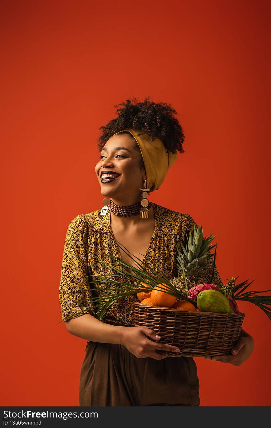 smiling young african american woman holding basket with exotic fruits on red,stock image