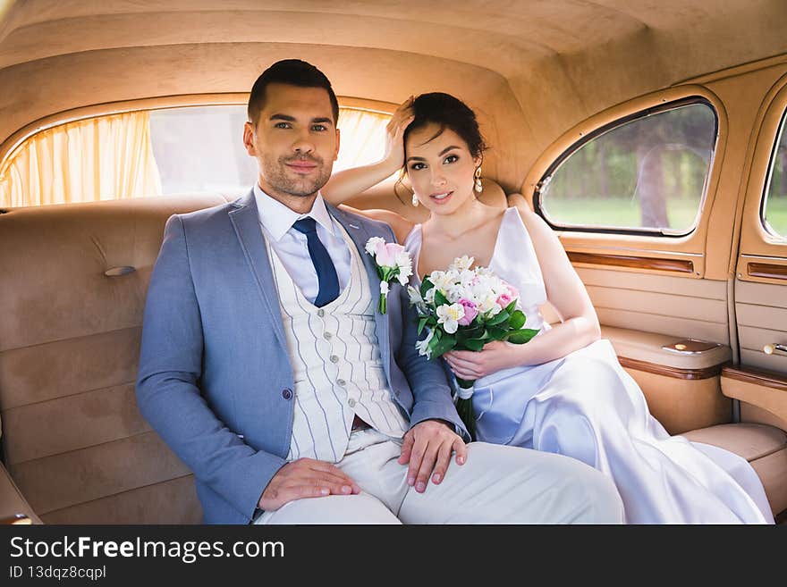 Smiling bride with bouquet looking at camera near groom in vintage car,stock image