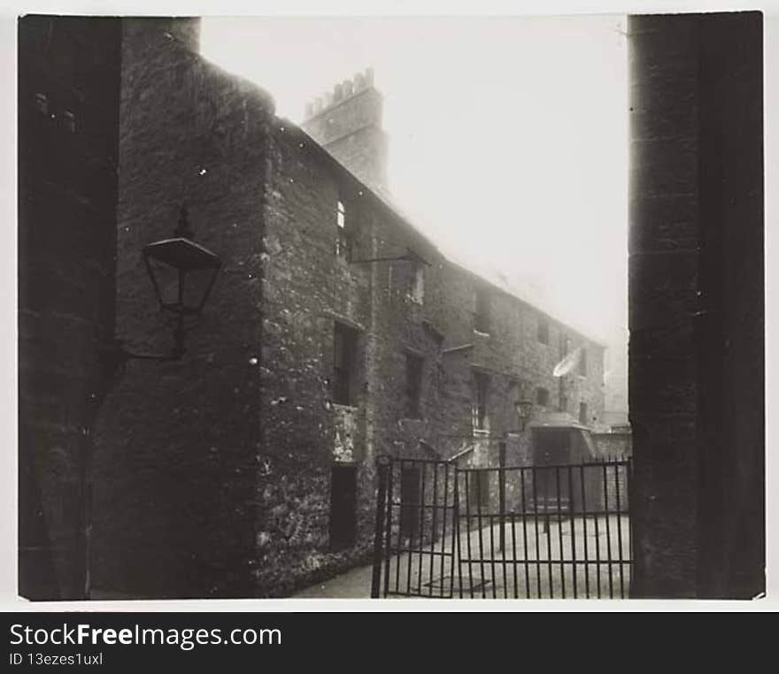 Photograph of a courtyard looking up to a two storey building . There are iron railings and a gate across the left side of the building and iron posts along the  front of the building. There is a street lamp on the left attached to the wall in the foreground. The  courtyard is concrete.

digital.nls.uk/74506932. Photograph of a courtyard looking up to a two storey building . There are iron railings and a gate across the left side of the building and iron posts along the  front of the building. There is a street lamp on the left attached to the wall in the foreground. The  courtyard is concrete.

digital.nls.uk/74506932