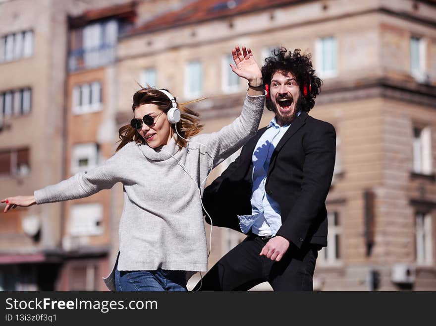 Young business colleagues dancing together with headphones after work. Happy friends jumping and fooling around outdoors.