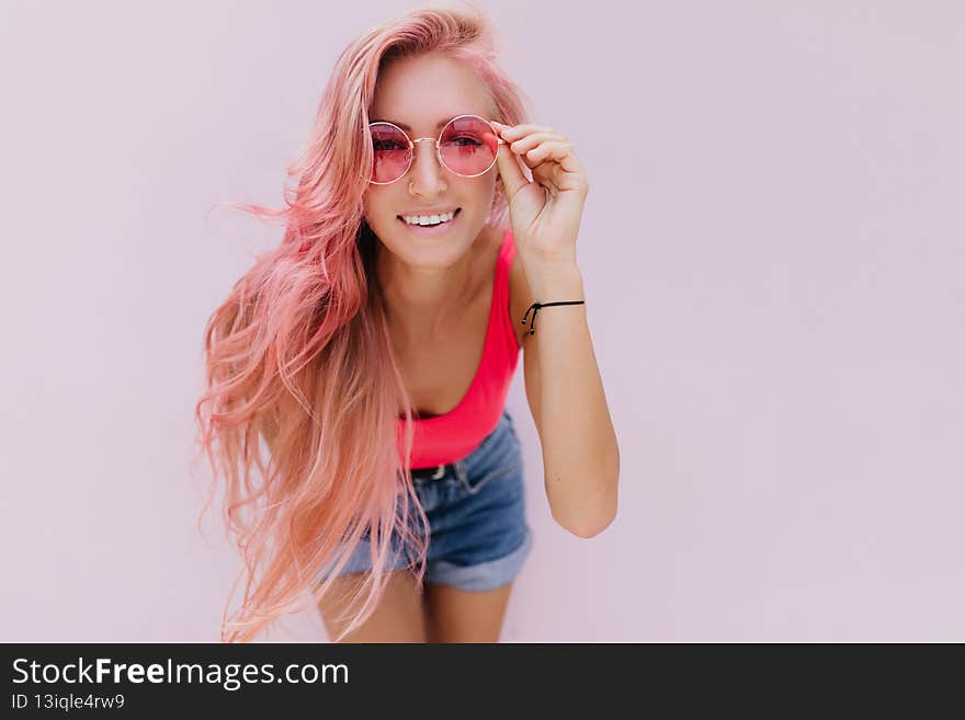 Interested girl with light-pink hair touching her sunglasses. Studio shot of attractive curly woman in trendy summer outfit smiling on white background